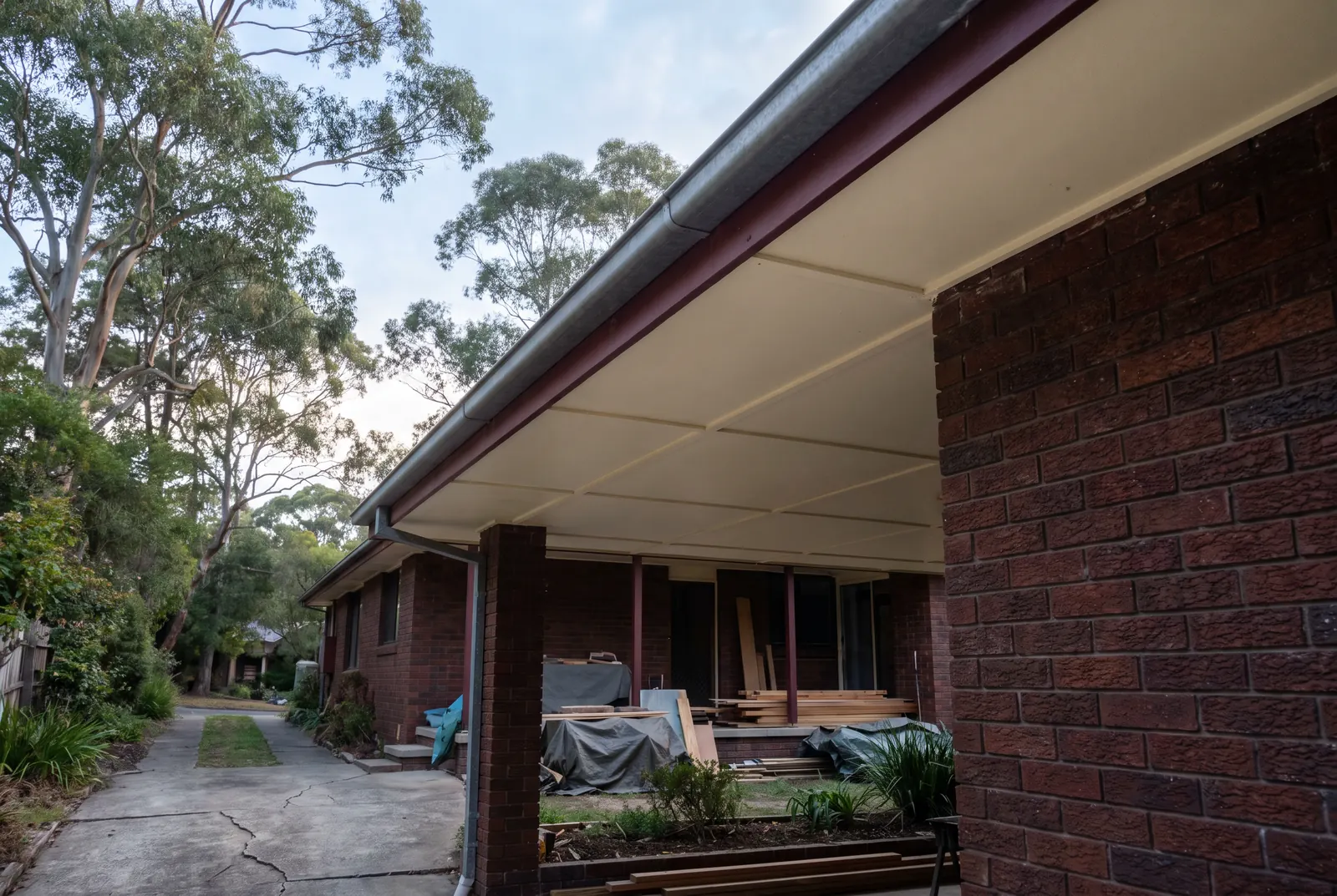 The underside of a brick house's eaves, lined with flat cream sheeting behind a red-brown fascia, with timber and tarps from a renovation on the patio below