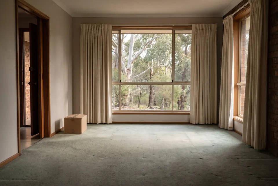 An empty living room after a clearance: bare carpet with faint marks where furniture stood, curtains still hanging, one cardboard box by the door and gum trees through a wide window
