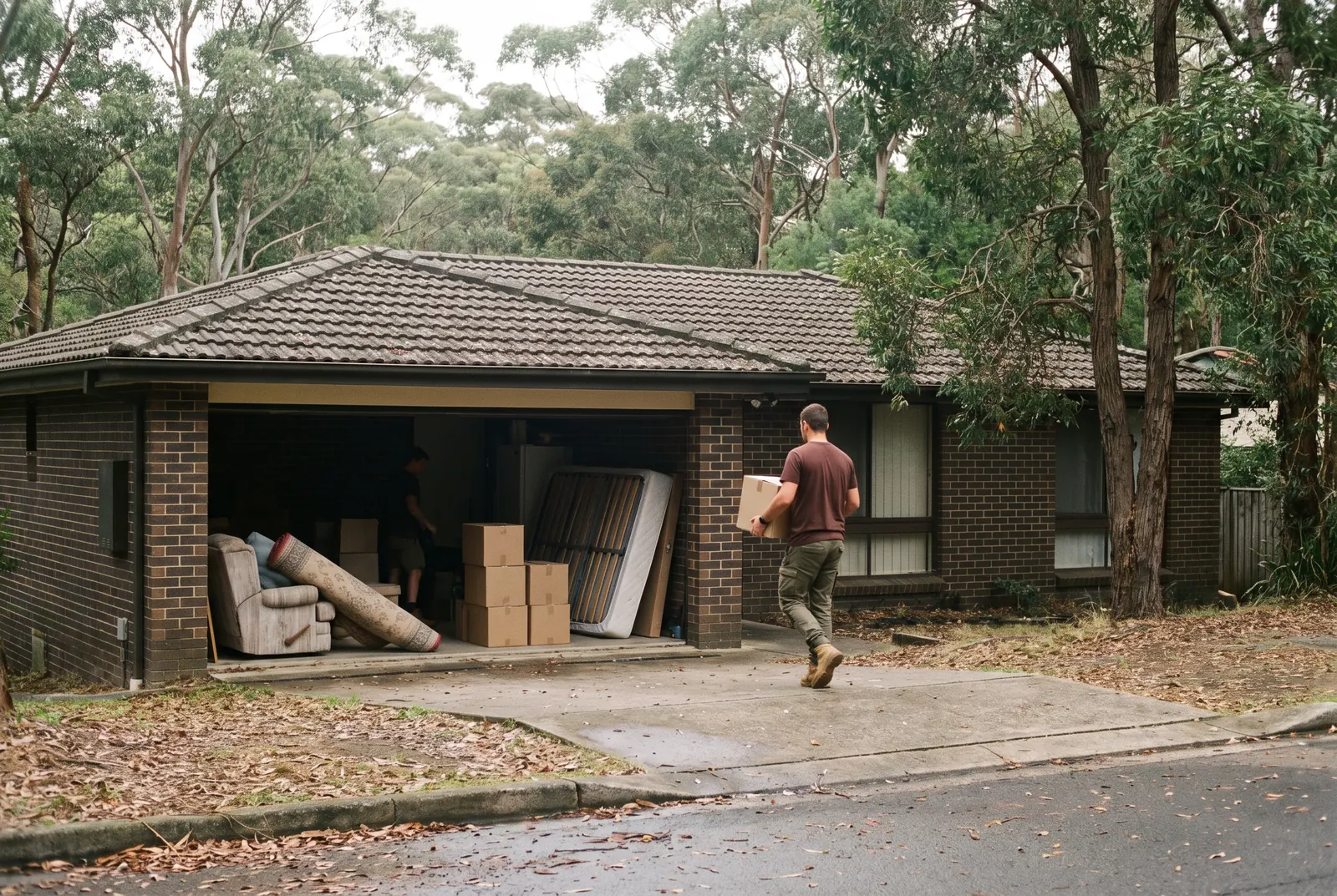 A single-storey brick house with its double garage open, an armchair, a rolled rug, a bed base and boxes waiting inside, and a crew member carrying a box down the drive
