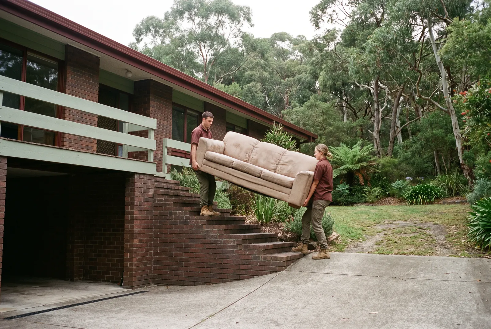 Two crew members carrying an old beige three-seater lounge on the outdoor brick steps beside a split-level house, the under-house garage behind them