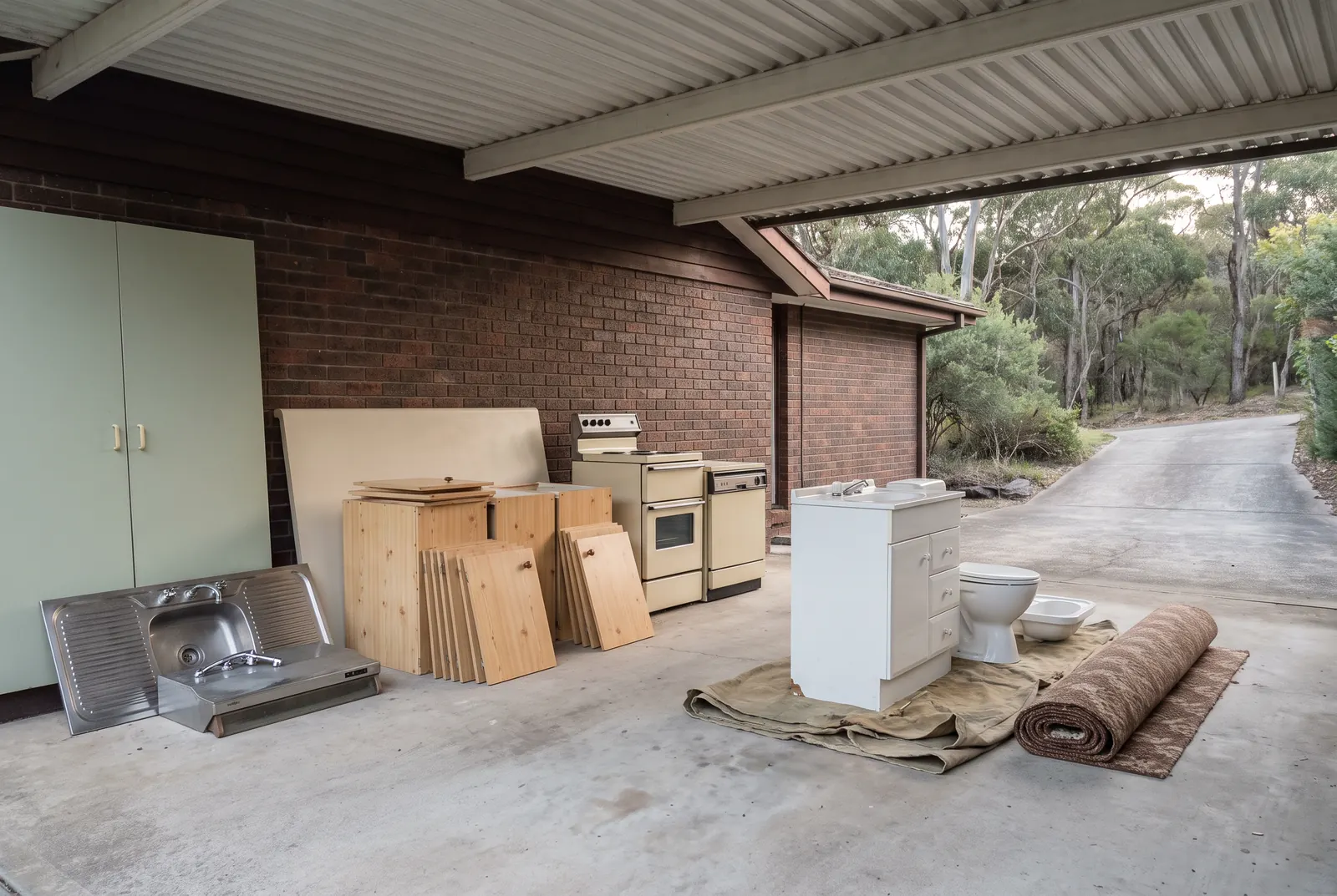 An old kitchen and bathroom sorted into groups under a carport: a stainless sink and rangehood, pine cabinets with their doors stacked flat, a benchtop on its edge, an oven and dishwasher, a vanity, toilet and basin on a tarp, and a rolled carpet