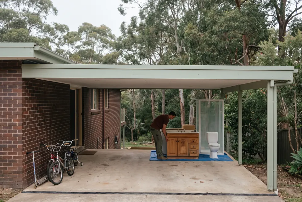 A carport beside a brick house backing onto bushland. An old timber vanity, a toilet and a shower screen wait on a blue tarp at the far end while a crew member lifts the vanity, and the children's bikes and scooter stand against the wall at the near end