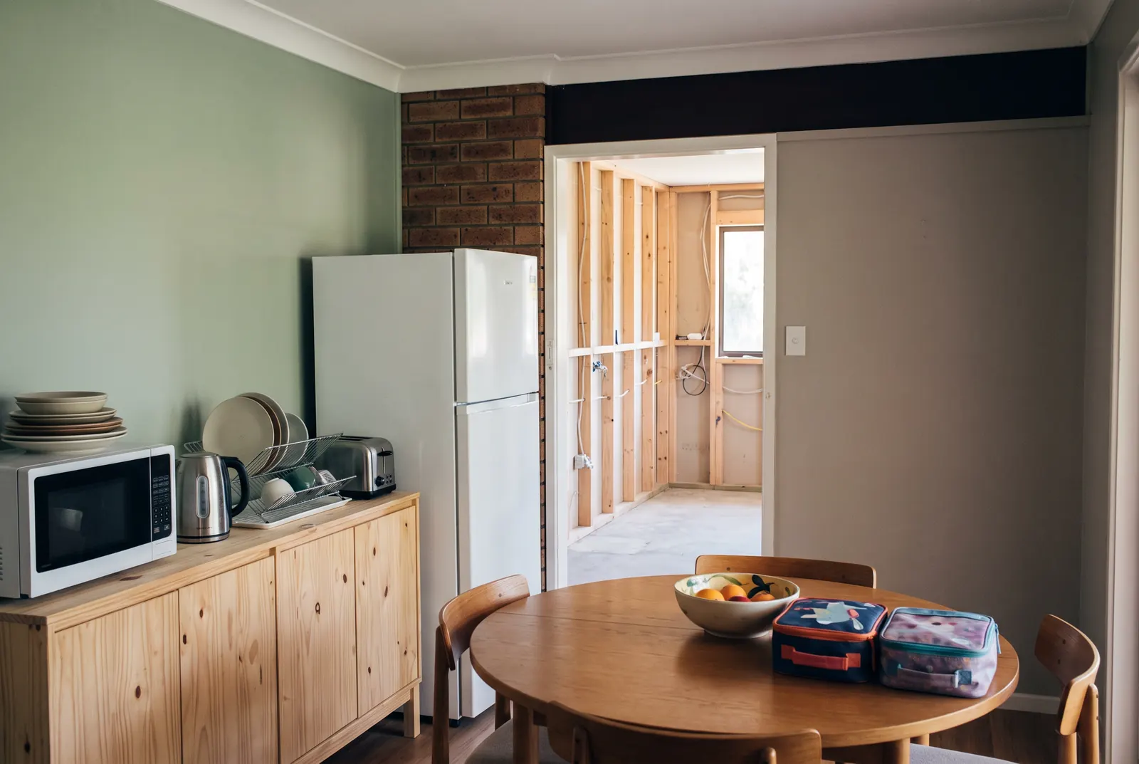 A dining room set up as a stand-in kitchen, with the fridge, a microwave, a kettle and a dish rack on a pine sideboard and school lunchboxes on the table. Through the doorway, the old kitchen is stripped to bare stud framing
