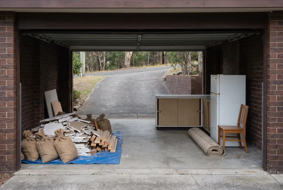 An under-house garage mid-renovation: bagged plaster, broken board and timber offcuts on a tarp on one side, and an old kitchen cabinet, a fridge, a chair and a rolled carpet set apart whole on the other