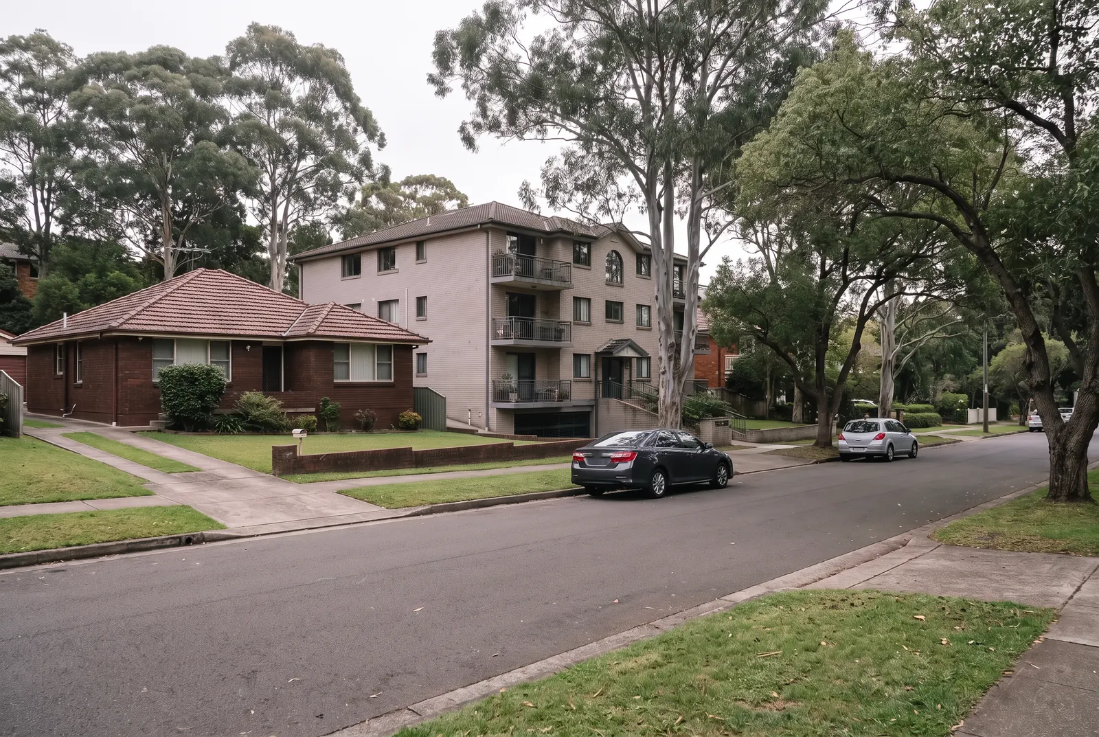 A leafy street with a single-storey brick house beside a three-storey brick apartment block and its car park entry, tall gums over the nature strip